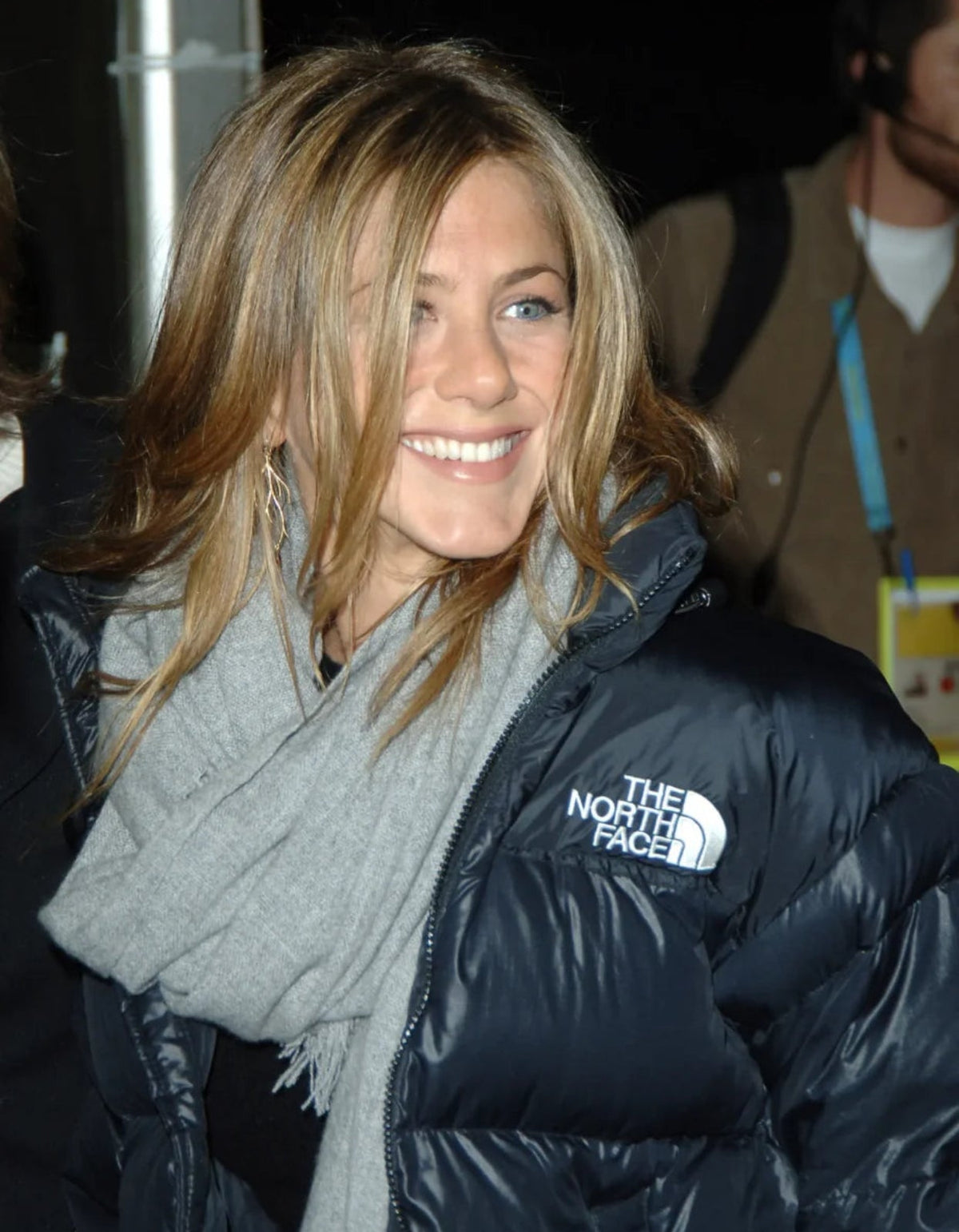 Jennifer Aniston smiling while wearing a black puffer jacket with a white The North Face logo and a light gray scarf.